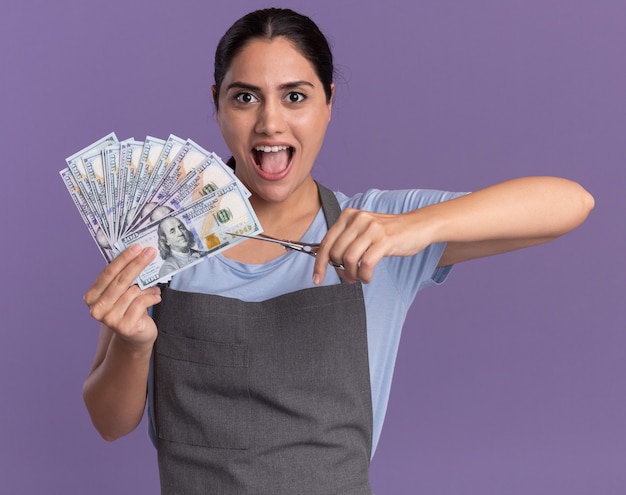 Happy young beautiful woman hairdresser in apron holding cash going to cut money with scissors emotional and excited looking at front standing over purple wall