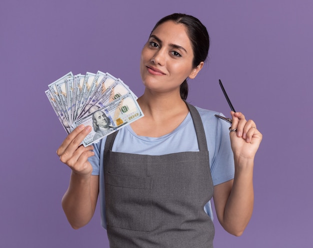 Happy young beautiful woman hairdresser in apron holding cash and scissors looking at front with smile on face standing over purple wall