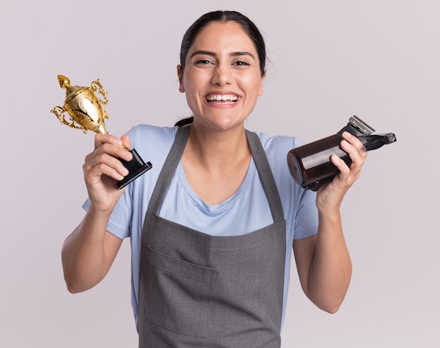 Happy young beautiful woman hairdresser in apron holding gold trophy trimmer machine with spray bottle looking at front with smile on face standing over white wall