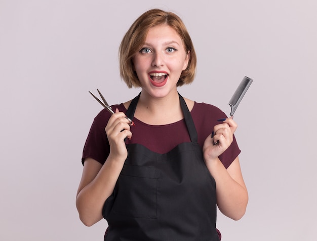 Happy young beautiful woman hairdresser in apron holding hair comb and scissors looking at front smiling standing over white wall