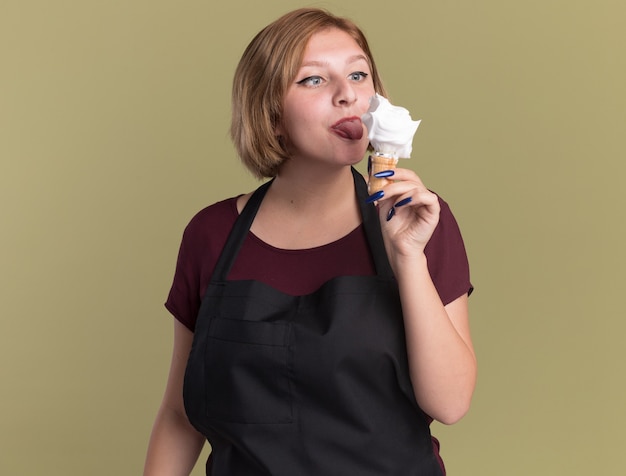 Happy young beautiful woman hairdresser in apron holding shaving brush with shaving foam looking at it imitating an icecream standing over green wall