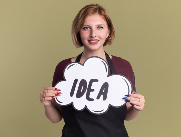 Happy young beautiful woman hairdresser in apron with scissors showing speech bubble sign with word idea looking at front with smile on face standing over green wall