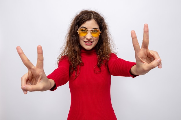 Free photo happy young beautiful woman in red turtleneck wearing yellow glasses smiling cheerfully showing v-sign standing on white