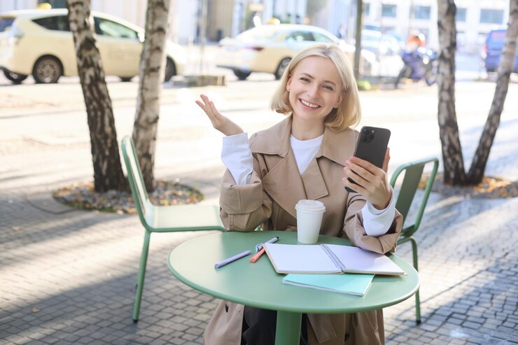 Free Photo happy young blond female model in coffee shop talks on video chat using smartphone and having