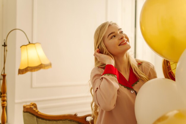 Happy young blonde caucasian girl looks away at party with balloons wears red nightgown Holiday concept