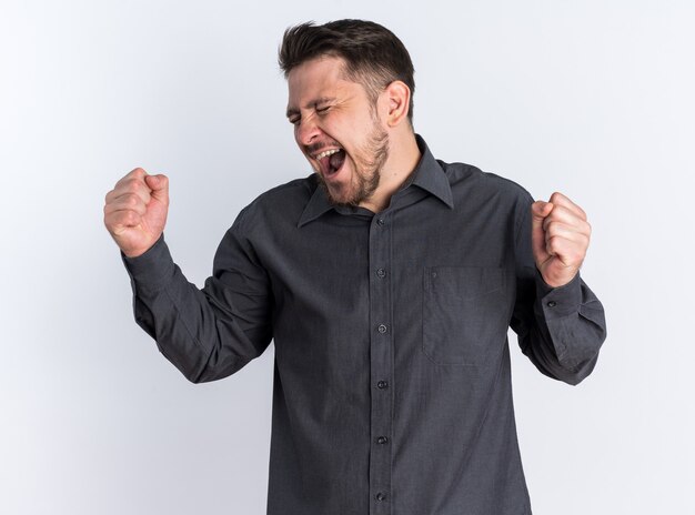 Happy young blonde handsome man doing yes gesture keeping fists up with open mouth and closed eyes 