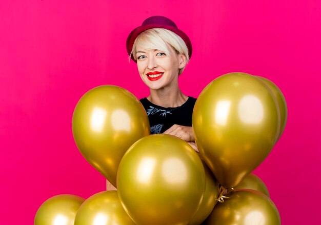 Happy young blonde party girl wearing party hat standing behind balloons looking at camera isolated on crimson background