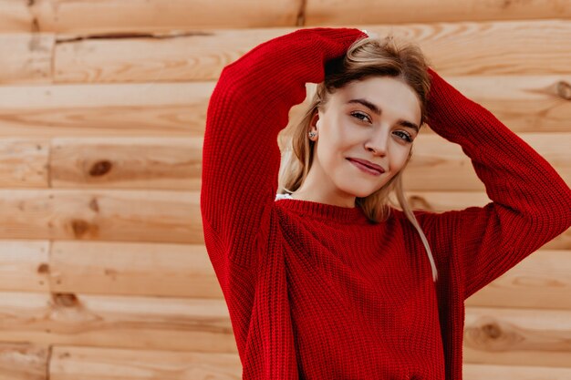 屋外の木製の壁にポーズをとってトレンディな赤いセーターで幸せな若いブロンド。笑顔のスタイリッシュな季節の服を着た美しい少女。