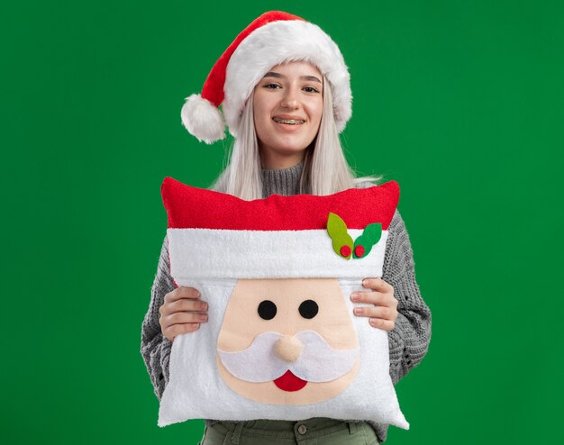 緑の背景の上に立っている顔に笑顔でカメラを見てクリスマス枕を保持している冬のセーターとサンタ帽子の幸せな若いブロンドの女性