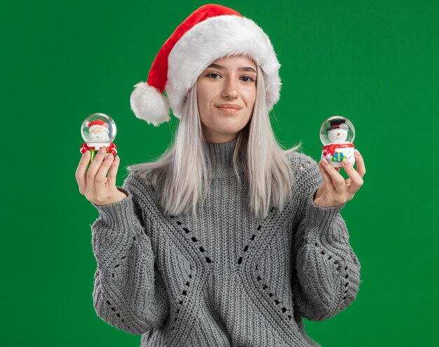 冬のセーターとサンタの帽子の幸せな若いブロンドの女性は、緑の背景の上に元気に立って笑顔のカメラを見てクリスマスのおもちゃのスノードームを保持しています
