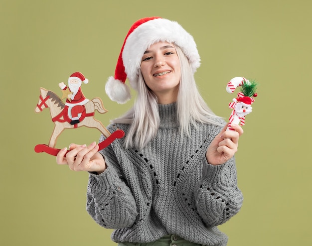 Happy young blonde woman in winter sweater and santa hat holding christmas toys  with smile on face  standing over green wall