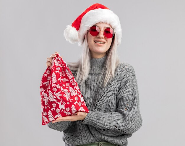 Happy young blonde woman in winter sweater and santa hat holding santa red bag with christmas gifts  smiling cheerfully standing over white wall