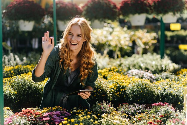 陽気な気分で幸せな若い植物学者の女の子は、花に囲まれたカーリーの女性研究者にサインOKを示しています