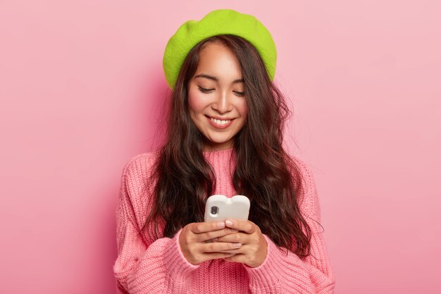 Happy young brunette Korean woman holds modern mobile phone, sends her photo to boyfriend, reads funny notification