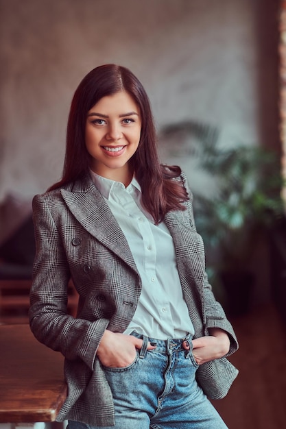Free photo happy young brunette woman dressed in a gray elegant jacket posing with hands in pockets while leaning on a table in a room with loft interior, looking at camera.