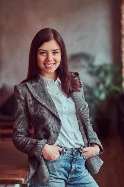 Happy young brunette woman dressed in a gray elegant jacket posing with hands in pockets while leaning on a table in a room with loft interior, looking at camera.