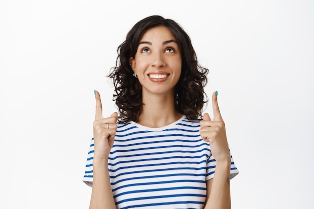Free photo happy young brunette woman looking and pointing up, smiling white teeth, standing cheerful against white background, wearing striped summer t-shirt