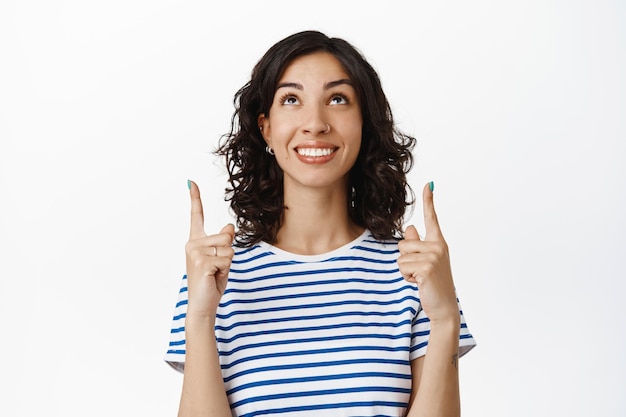 Happy young brunette woman looking and pointing up, smiling white teeth, standing cheerful against white background, wearing striped summer t-shirt