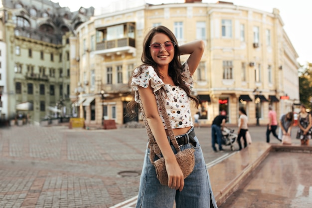Happy young brunette woman in loose stylish denim pants and cropped floral top smiles widely