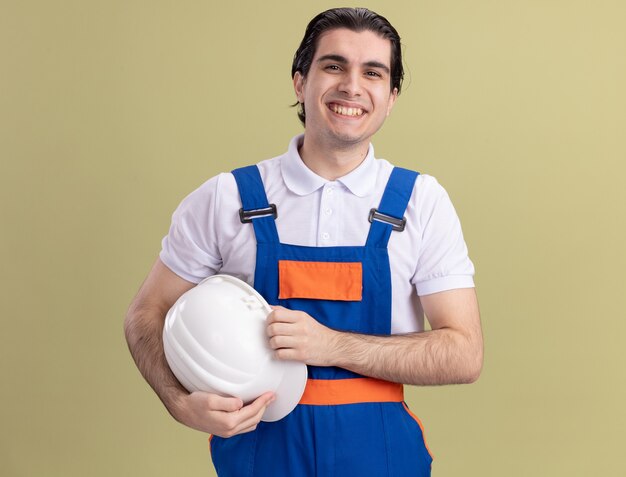 Счастливый молодой человек-строитель в строительной форме, держа свой защитный шлем, глядя вперед с улыбкой на лице, стоя над зеленой стеной