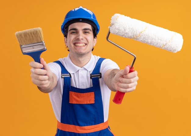 Free photo happy young builder man in construction uniform and safety helmet holding paint brush and roller looking at front smiling cheerfully standing over orange wall