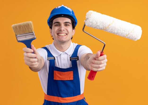 Happy young builder man in construction uniform and safety helmet holding paint brush and roller looking at front smiling cheerfully standing over orange wall