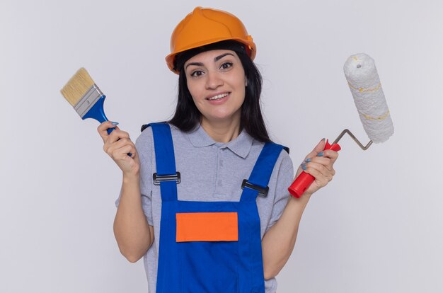 Happy young builder woman in construction uniform and safety helmet holding brush and paint roller looking at front samiling confident standing over white wall