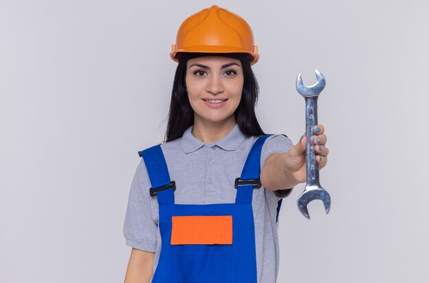 Happy young builder woman in construction uniform and safety helmet holding wrench looking at front smiling confident standing over white wall