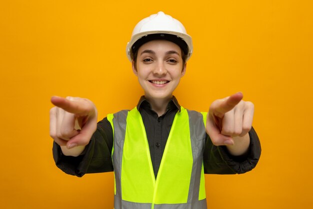 건설 조끼와 안전 헬멧을 쓴 행복한 젊은 건축업자 여성은 주황색 위에 검지 손가락으로 자신을 가리키며 웃고 있습니다.