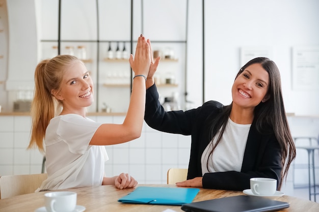 Happy young business women giving high five and celebrating success, sitting at table with documents and coffee cups, looking at camera