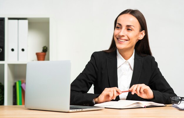 Happy young businesswoman sitting behind the desk with book; pen and laptop