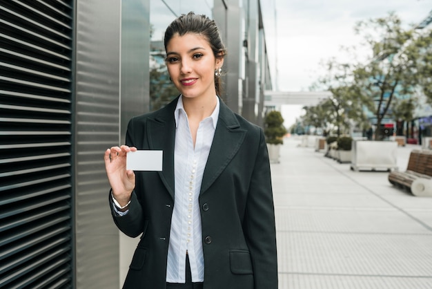 Happy young businesswoman standing outside the office building showing her business card