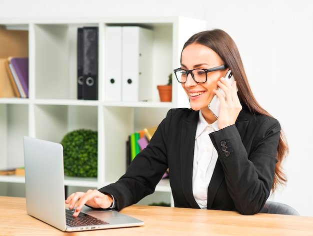 Happy young businesswoman talking on smartphone typing over laptop