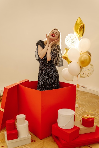 Happy young caucasian blonde woman in black dress stands in large box among gifts and balloons Entertainment and lifestyle concept