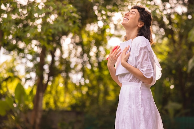 Happy young caucasian brunette woman closing her eyes is inspired by nature holding album and pencil in bunches outdoors