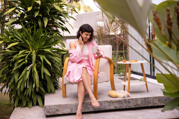 Happy young caucasian brunette woman in pink clothes with cup of coffee on vacation sits in courtyard of hotel Pleasure concept