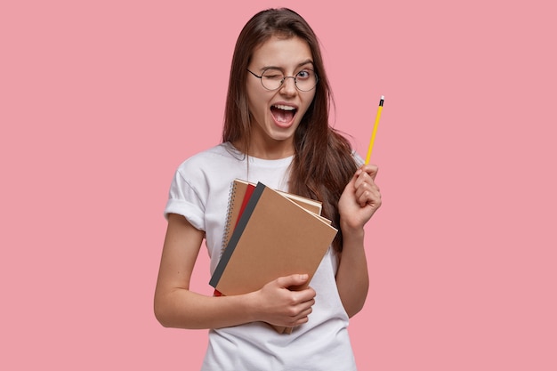 happy young Caucasian female genius gets good idea, blinks eye, holds pencil, carries notebooks, has fun indoor