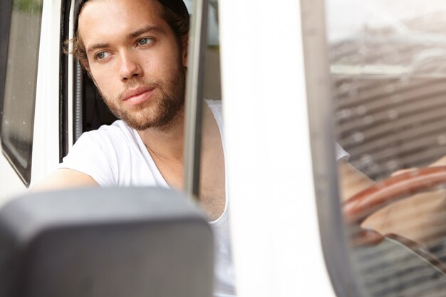 Happy young Caucasian hipster man wearing t-shirt driving his white car, smiling and looking out of open window, on his way home