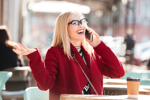 Happy young caucasian lady talking by phone drinking coffee.