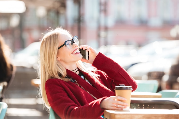 Happy young caucasian lady talking by phone drinking coffee.