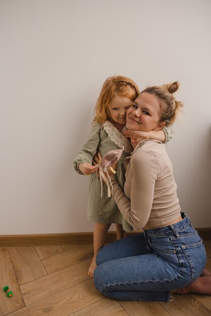 小さな赤毛の娘と幸せな若い白人の母親は部屋の床に座っている間カメラを見る母性の概念