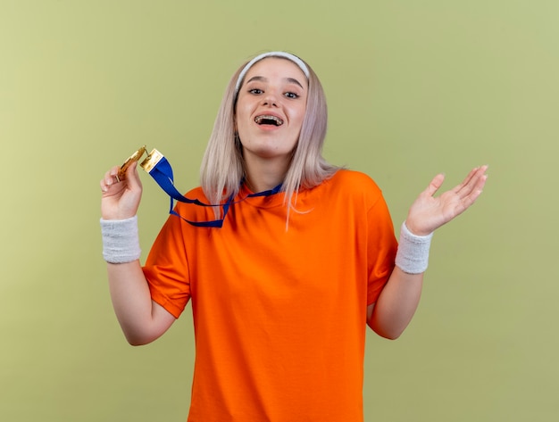 Happy young caucasian sporty girl with braces wearing headband and wristbands holds gold medal 