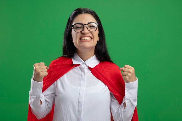 Happy young caucasian superhero girl wearing glasses smiling and clenching fists isolated on green wall with copy space