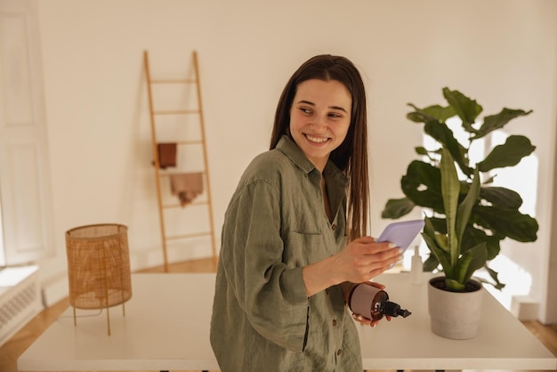 Happy young caucasian woman holding mobile phone looking away while standing in room Brunette girl reads love message in personal chat with smile on her face Concept of enjoying moment