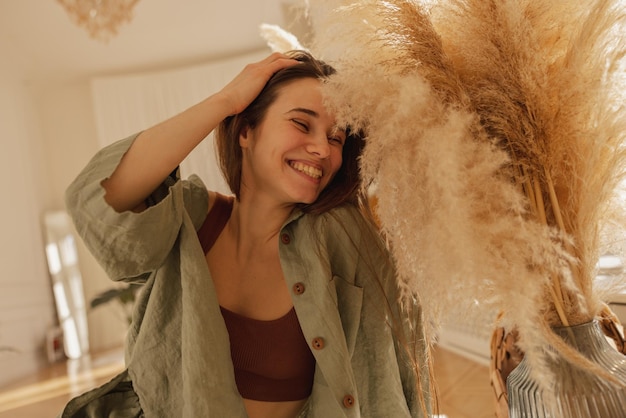 Free photo happy young caucasian woman smiles broadly with eyes closed while sitting on floor in light room brunette fixing her hair and wears casual clothes concept of enjoying moment