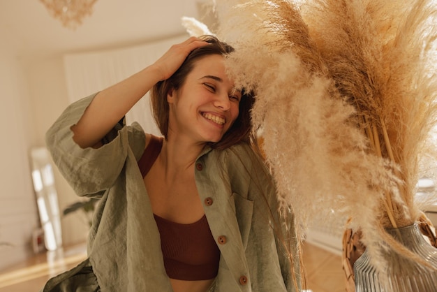 Счастливая молодая кавказская женщина широко улыбается с закрытыми глазами, сидя на полу в светлой комнате брюнетка поправляет волосы и носит повседневную одежду концепция наслаждения моментом Бесплатные Фотографии