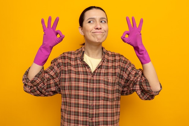 Счастливая молодая уборщица в повседневной одежде в резиновых перчатках, весело улыбаясь, показывая знак ОК, стоящий над оранжевой стеной