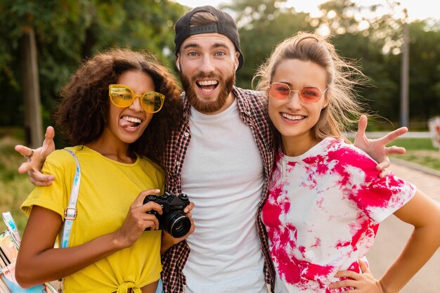 Счастливая молодая компания эмоционально улыбающихся друзей, гуляющих в парке с фотоаппаратом, мужчины и женщины, весело проводящие время вместе