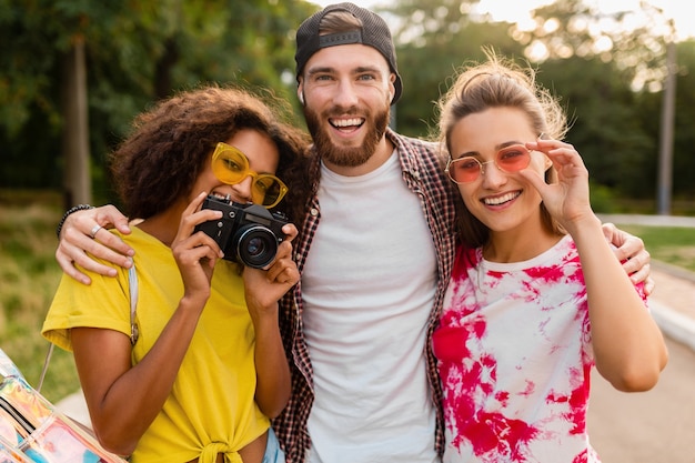 Happy young company of emotional smiling friends walking in park with photo camera, man and women having fun together