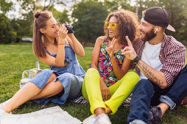 公園に座っている友人の幸せな若い会社、一緒に楽しんでいる男性と女性、カラフルな夏の流行に敏感なファッションスタイル、カメラで写真を撮る旅行、話す、笑顔